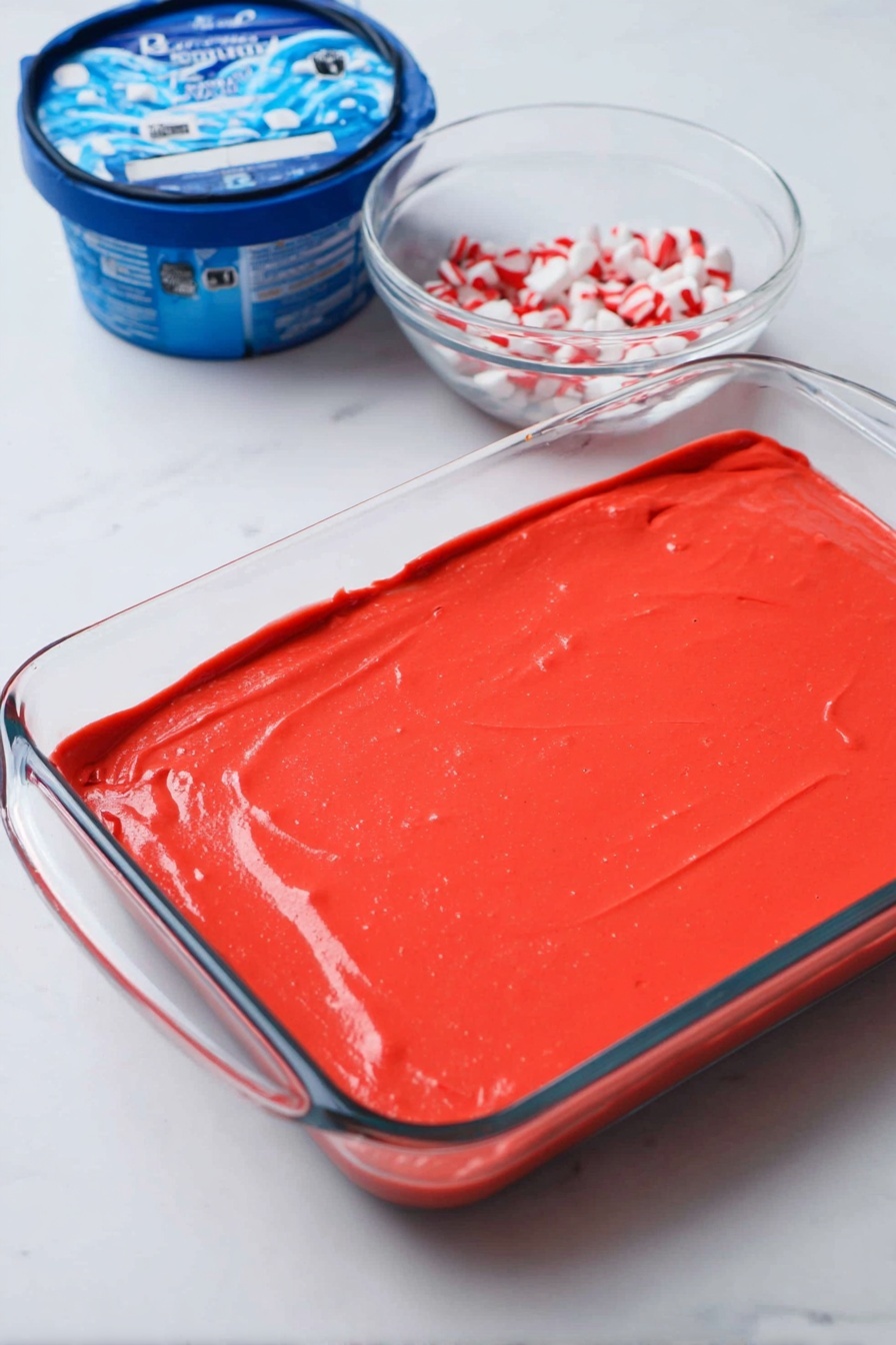 A slice of dessert with three visible layers sits on a white plate over a white marbled surface. The bottom layer is dark brown, crumbly, and looks like crushed cookies or chocolate crumbs. The middle layer is thick, creamy white with a smooth texture. The top layer is white whipped cream sprinkled with small, bright red and white candy pieces. A fork is holding a piece of the dessert on the right side, showing the inside layers clearly. In the background, a red and white striped cloth can be seen. photo taken with an iphone --ar 2:3 --v 7